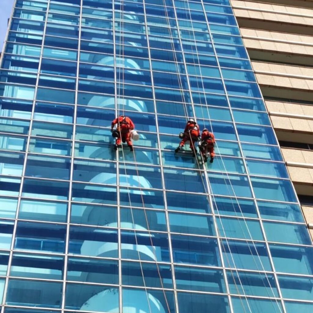 Três profissionais da Vetor Soluções executando manutenção e limpeza em fachada de vidro corporativa utilizando técnicas de alpinismo industrial, com rigoroso cumprimento da NR-35.