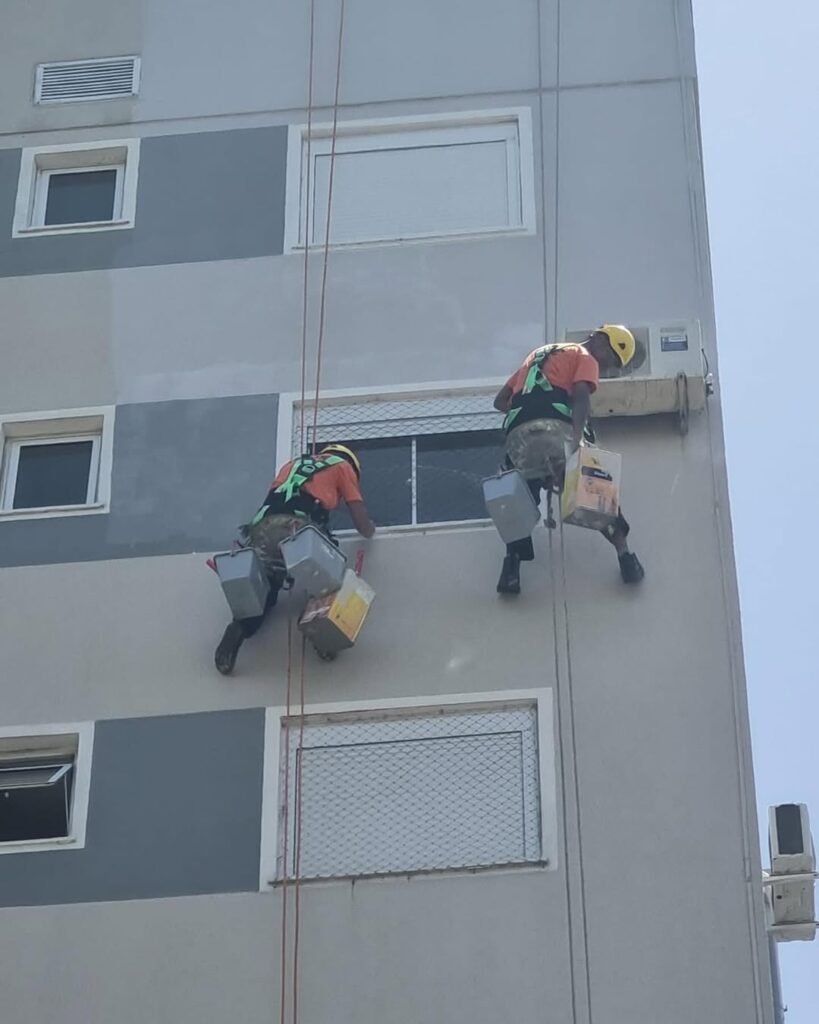 Dois profissionais da Vetor Soluções realizando pintura e revitalização de fachada predial com técnica de alpinismo industrial, garantindo segurança e responsabilidade técnica (ART).
